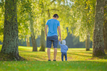 Young adult father and baby boy walking on green grass through tree alley at park. Spending time together in beautiful autumn day. Back view. Lovely emotional moment. Peaceful atmosphere in nature.