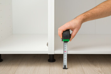 Young adult man hand holding measure tape and measuring height between floor and white base cabinet...