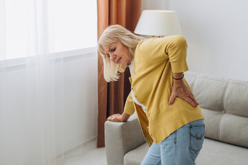 Senior woman having backache and touching her back in living room at home