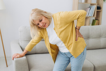Senior woman having backache and touching her back in living room at home