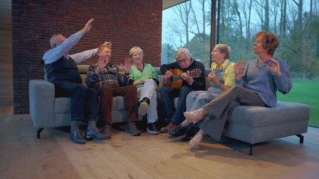 group of senior friends sitting on sofa playing guitar and singing songs together