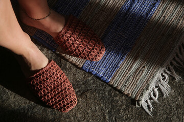 Woman standing in handmade raffia mules on the striped handwoven wool rug. Sustainable ethically made pair of slippers. Responsibly sourced shoes with eco-friendly materials. - obrazy, fototapety, plakaty