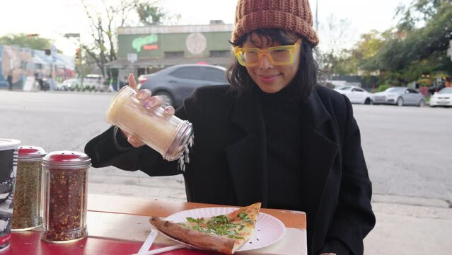 Cute Asian woman shakes parmesan topping onto margherita pizza slice at outdoor street pizza restaurant. slow motion