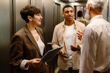 Three diverse colleagues smiling while discussing business issues in elevator