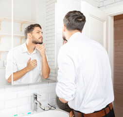 Obraz premium Handsome businessman looking at mirror and applying moisturizing cream on cheeks in bathroom. Groomed young guy doing skincare morning routine after taking a shower