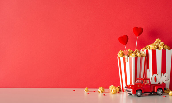 Valentine's Day film debut ambiance: Side view of table with small car denoting food delivery, striped boxes brimming with popcorn, heart decorations, sprinkles against red wall. Ad space available