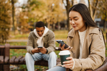 Beautiful couple drinking coffee and using smartphones while spending time together in park