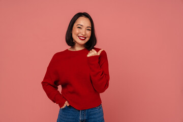 Smiling asian woman showing thumb up while standing isolated over pink background