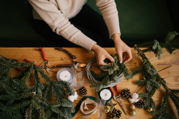 Woman making Christmas wreath using natural pine branches, sitting near wooden table. © polinaloves