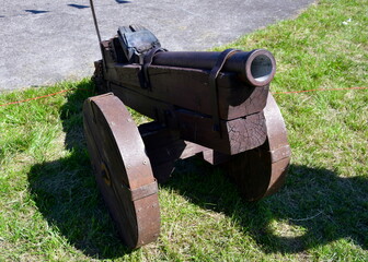 A close up on a wooden bombard or cannon made out of planks, boards, and logs with some iron elements attached to it seen on a sunny summer day next to some field, shrubs, and concrete path