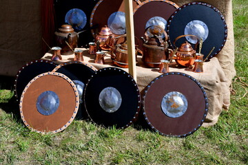 A close up on a medieval encampment with various historic pieces of equipment, including shields, helmets, metal tools, vases, cups, containers, leather jackets, and elements of armour seen in summer