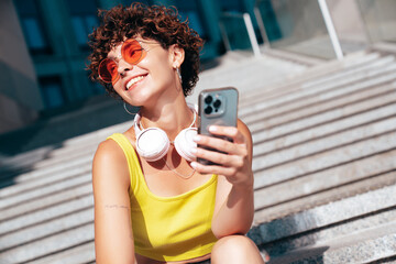 Young beautiful smiling hipster woman in trendy summer clothes. Carefree woman with curls hairstyle, in the street. Positive model holds mobile phone, looks at cellphone screen, uses smartphone apps