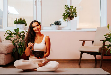 Happy beautiful young woman is sitting in lotus position and palyying singing bowl on yoga mat at...