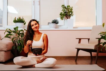 Foto op Aluminium Happy beautiful young woman is sitting in lotus position and palyying singing bowl on yoga mat at yoga class. Soundhealing concept. © dvulikaia