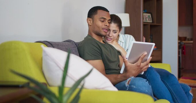 Happy young millennial couple looking at tablet computer on the couch together and laughing. Attractive African American and Caucasian couple using mobile device and smiling. 4k slow motion handheld