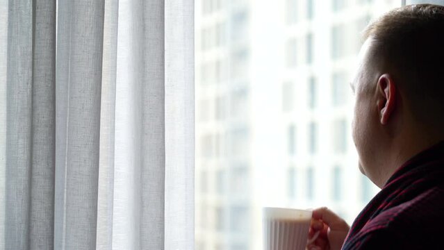 Mature man in bathrobe opens curtains to meet new day, drinks coffee and looks out window in morning