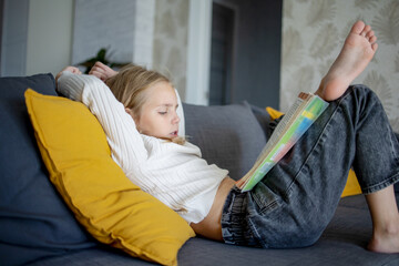 At home, a charming girl with blonde hair is sitting on the couch, and she is reading an exciting book.