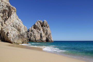 Fototapeta na wymiar The bright rocks are located on the clear sandy shore of the ocean with water turquoise color. Pacific Ocean; Cabo San Lucas, Mexico.