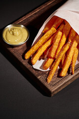 vertical shot of crispy French fries inside of paper cone near dipping sauce on wooden cutting board