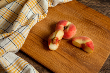 Cutting board with whole and half of saturn peaches or flat peaches on wooden background with yellow kitchen towel..