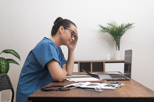 tired exhausted doctor in blu uniform sitting in her offie near laptop with heap of patients tes results and medical documents papers
