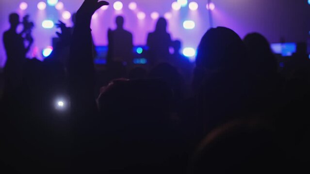Silhouettes of people jumping, waving, taking photos or recording videos of a live music concert on smartphones at an open-air festival. Bright colorful stage lighting.  