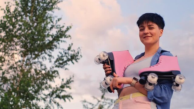 A young smiling girl with short hair holding roller skates in her hands. Woman in the park with rollerblades. first steps and summer sports training concept.