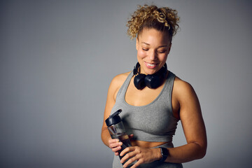 Studio Shot Of Woman In Gym Fitness Clothing With Wireless Headphones And Water Bottle On Grey