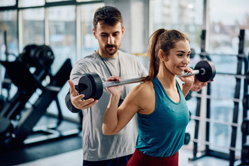 Happy athletic woman exercising with barbell with coach's assistance in gym.