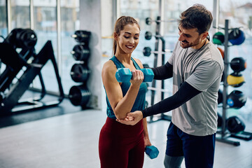 Happy athletic woman exercising with dumbbells while fitness instructor is assisting her in health club.