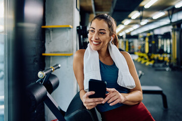 Happy athletic woman listens music over earbuds while using cell phone in gym.
