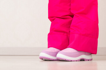 Little girl legs in pink warm waterproof ski trousers and boots standing on wooden home floor....