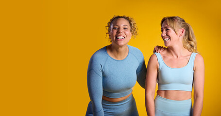Studio Portrait Of Two Smiling Women Wearing Gym Fitness Clothing Exercising On Yellow Background