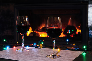 Wine and fireplace. Two glasses of red wine against the backdrop of a burning fireplace and New Year’s garlands. Dark romantic relaxation scene.