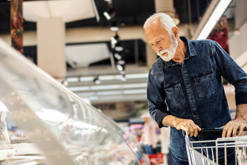 Portrait of mature Caucasian man doing grocery shopping at the supermarket. Smiling man pushing a...