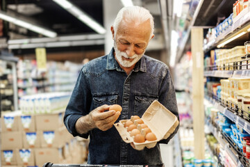 A old Caucasian businessman is standing in the refrigerated section of a supermarket and picking...