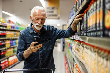 Senior Man doing grocery shopping at the supermarket, he is using apps on his smartphone. Male...