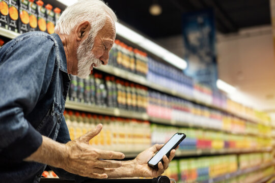Mature man shopping healthy food in supermarket blur background. Close up view man buy products using cellphone in store. Person comparing the price of produce.