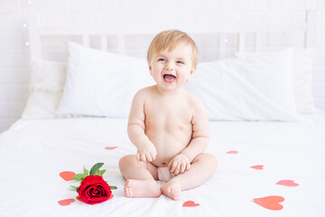 laughing baby boy blonde is sitting on the bed in diapers and with a red rose at home and smiling...
