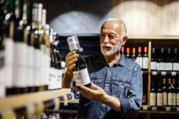 Male consumer buying bottle of red wine in winery store. Smiling mature man wearing casual clothes...