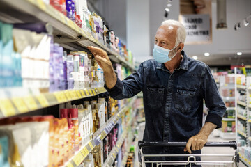 Senior man wearing protective face mask, choosing cosmetic products at supermarket, copy space.