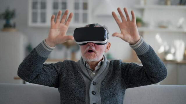 Senior man playing in virtual glasses. Happy amazed male in electronic goggles watching movie playing video games sitting on couch at home. Enjoy online world in VR helmet. Cyberspace, entertainment.