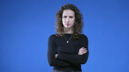 Upset woman crossing arms looking at camera while standing on blue background. Angry female 20s...