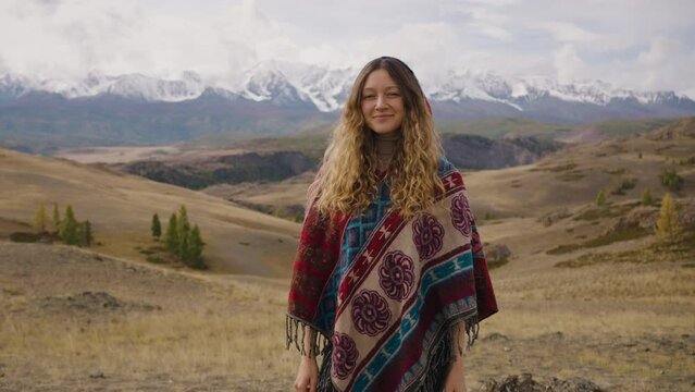 Portrait of a female traveler standing against the background of snowy mountains in a poncho looks at the camera and smiles