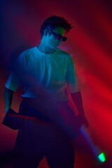 Handsome stylish young guy in white t-shirt and sunglasses posing over red background in neon lights. Night club. Concept of youth culture, fashion and male beauty, emotions, inspiration, trends