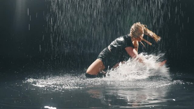 Sensual woman enjoy dancing under rain. Splashing sitting in puddle in black shorts and top. Beautiful installation performance of blonde girl. Emotional sensual movement of sexy dancer in rainy room.