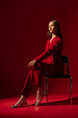 Fashionable confident woman wearing trendy red suit blazer, classic trousers, metallic silver color shoes, sitting on chair, posing on red background. Full-length studio fashion portrait
