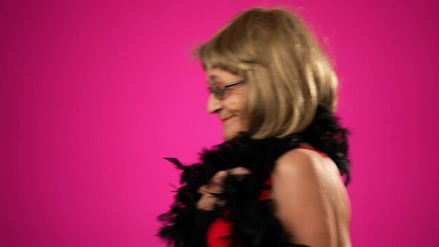 Funny elderly woman walks across frame throws boa over shoulder, and smiles at camera. Portrait of old woman isolated on pink background