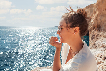 young latino woman in white summer cloths sitting at a cliff at Algarve coast in Portugal, looking...