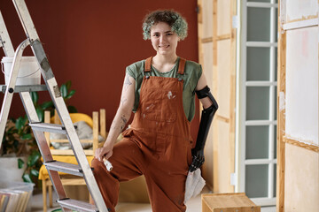 Portrait of young painter smiling at camera while using ladder to paint wall in the room